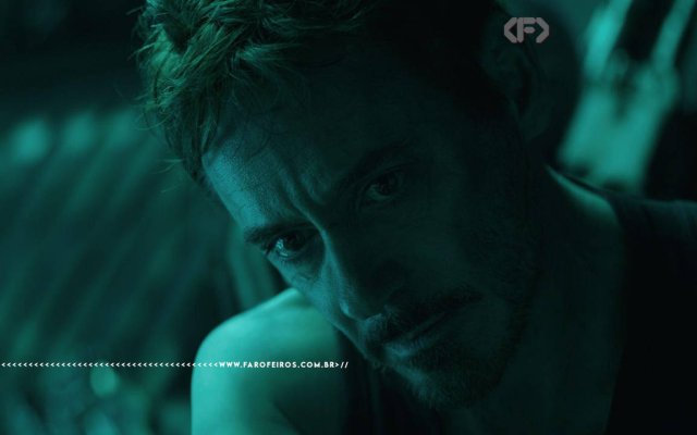 Cena do filme Vingadores: Ultimato com iluminação esverdeada. Close no rosto de Tony Stark, interpretado por Robert Downey Jr., com expressão abatida e exausta, olhando para baixo. Na parte inferior, a marca d'água do site www.farofeiros.com.br