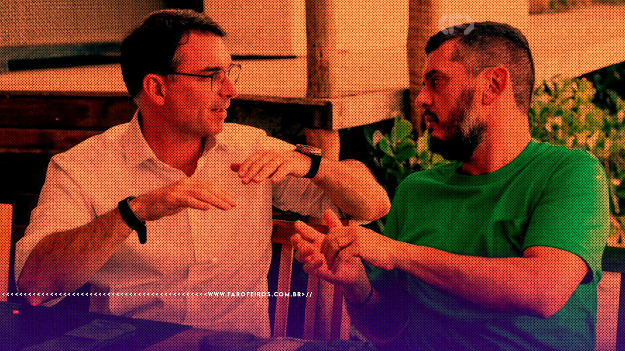 Fotografia com filtro laranja e roxo mostrando Flávio Bolsonaro, de camisa social clara e óculos, conversando e apertando a mão de Rodrigo Bacellar, que veste camiseta verde, em um ambiente informal.