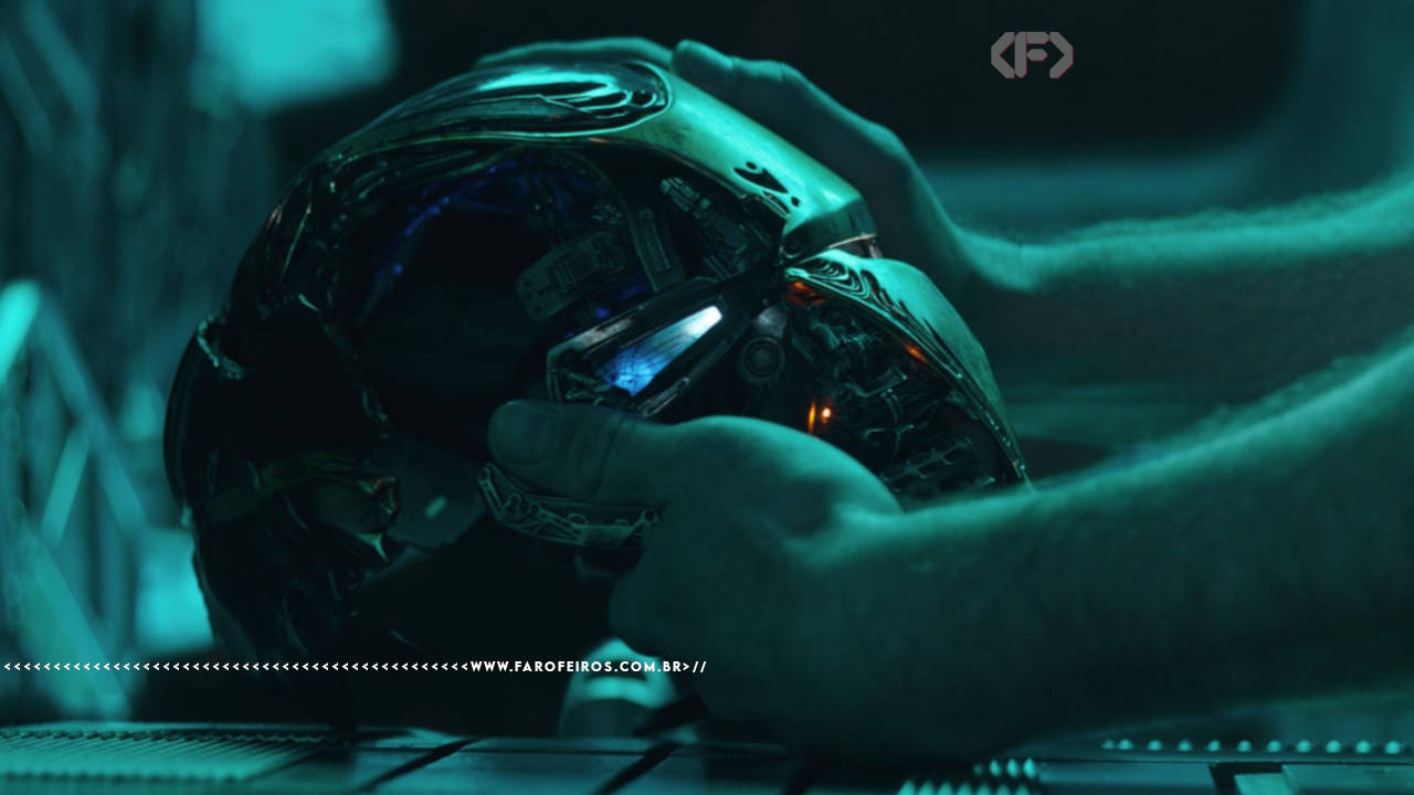 Cena do filme Vingadores: Ultimato com iluminação esverdeada. Em close, as mãos de Tony Stark seguram o capacete da armadura do Homem de Ferro, com os visores iluminados em azul. A peça exibe detalhes metálicos intrincados e danificados. Na parte inferior, a marca d'água do site www.farofeiros.com.br