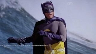 Batman de Adam West surfando de sunga amarela em um corte de cena da cintura para cima e braços jogados para a esquerda.